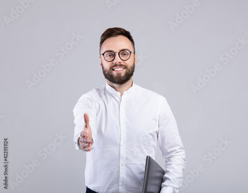 Bearded young businessman with laptop computer offering hand for handshake on grey studio background © Prostock-studio
