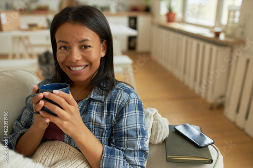 Фотография Happy young black woman in checkered shirt sitting on sofa with mug in her hands, drinking warm herbal tea, recovering from cold under blanket, reading book or using cell phone