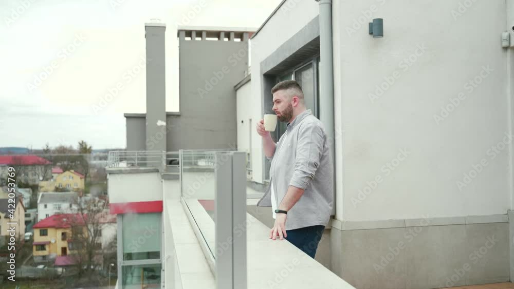 young handsome bearded man standing on modern balcony or terrace enjoying city view on urban background. business person or freelancer casual clothes drinking a hot coffee or tea at home or office young handsome bearded man standing on modern balcony or terrace enjoying city view on urban background. business person or freelancer casual clothes drinking a hot coffee or tea at home or office