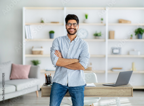 Happy freelance arab guy standing at desk at home office, posing and smiling to camera