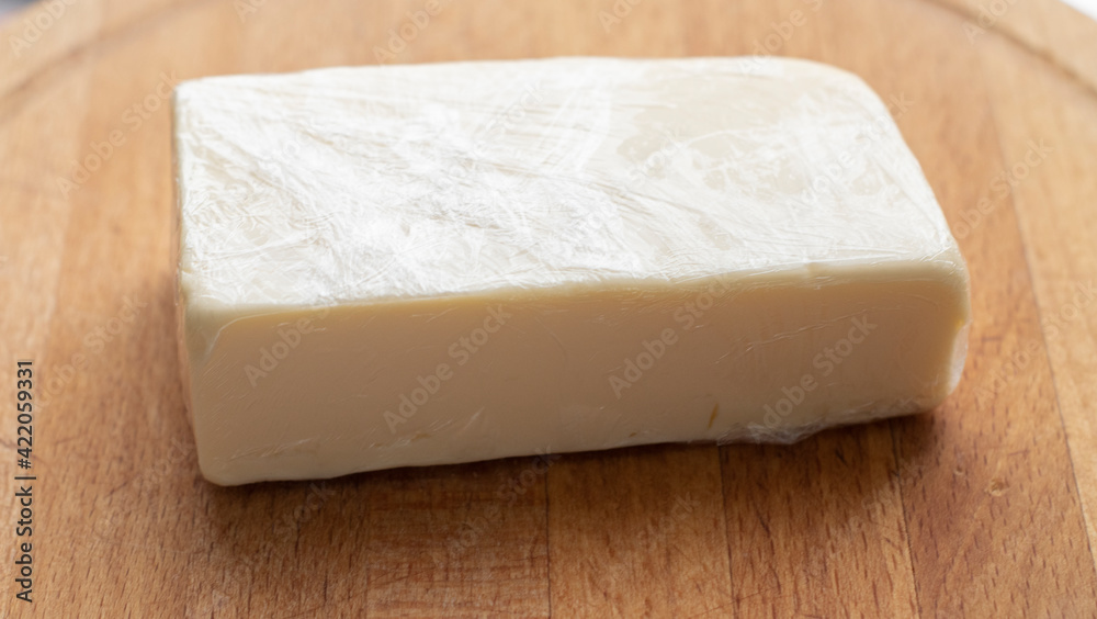 a piece of cheese in a plastic wrap lies on a wooden board, on a light background. Food, lactose-free cheese, healthy food, daily diet