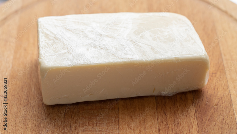 a piece of cheese in a plastic wrap lies on a wooden board, on a light background. Food, lactose-free cheese, healthy food, daily diet a piece of cheese in a plastic wrap lies on a wooden board, on a light background. Food, lactose-free cheese, healthy food, daily diet