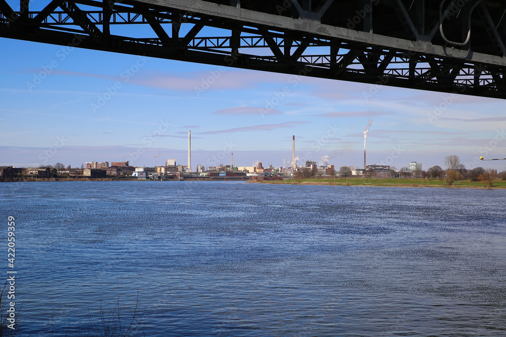 Fototapeta premium Krefeld (Uerdingen) - March 1. 2021: View beyond steel bridge over river rhine on industrial area with factories and chimneys against blue sky