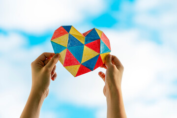 Child hands holding colorful heart over blue sky background. World autism awareness day concept