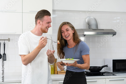 young caucasian boyfriend feeding organic healthy fruit salad for breakfast to girlfriend in kitchen. couple together concept