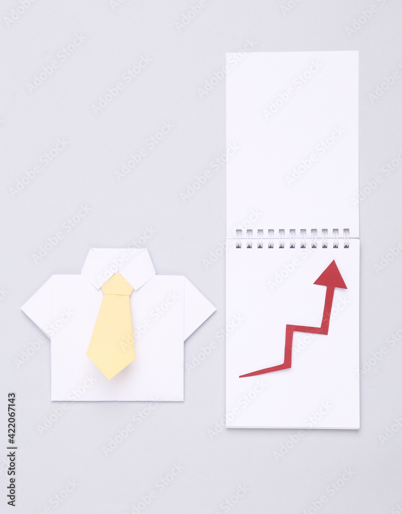 Notepad with growth arrow and origami shirt on gray background. Business concept. Level up Notepad with growth arrow and origami shirt on gray background. Business concept. Level up