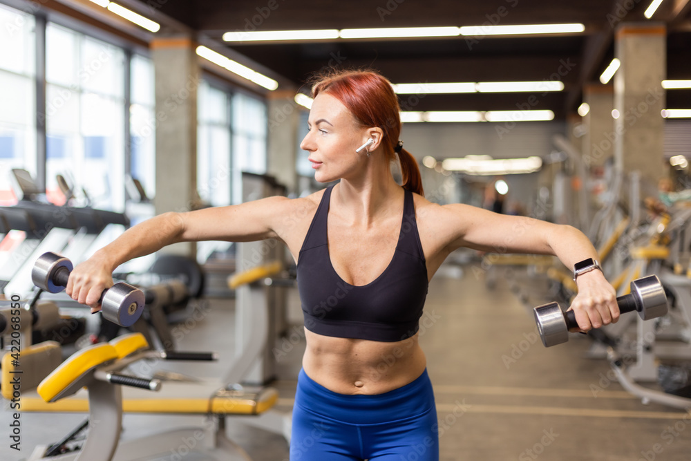 Attractive red-haired fitness woman trains shoulder muscles with dumbbells in a modern gym. Bodybuilding and Fitness