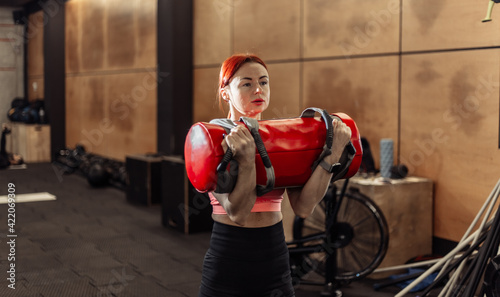 Foto Muscular woman is practicing intense workout with sand bag in the gym