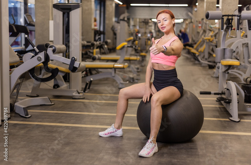 Red-haired cheerful fit woman sits on a fitball in a modern gym. Shows thumb up © splitov27