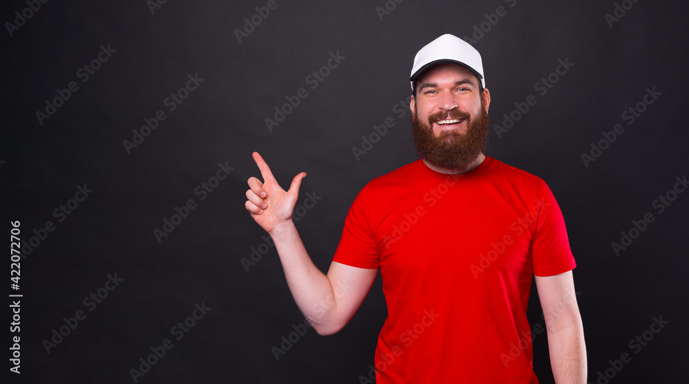 Happy handsome young bearded man in red t-shirt pointing at the corner over black background Happy handsome young bearded man in red t-shirt pointing at the corner over black background