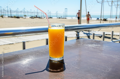 Glass with plastic straw filled with freshly squeezed orange juicestands on table of beach cafe against backdrop of the beach and sea. Beach vacation concept, healthy drinks.