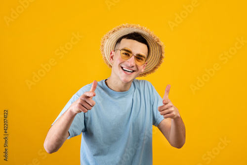 Portrait of handsome Caucasian man in straw hat and sunglasses pointing at camera on orange studio background