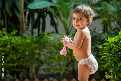adorable toddler stands with a toy in his hands against a background of greenery