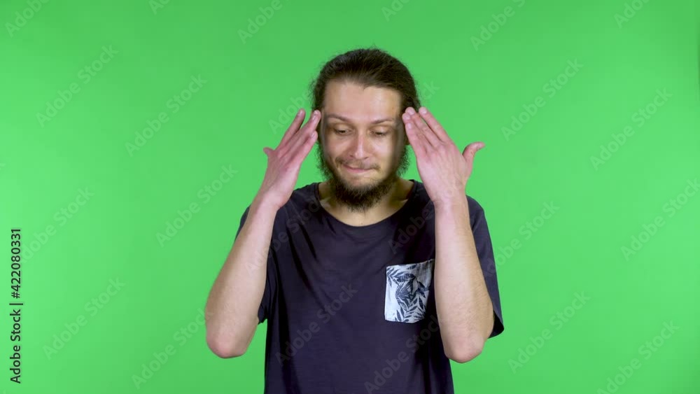 Portrait of a young man looking at the camera, shrugs his shoulders, puts his hands to his temples and says something smiling. Bearded darkhaired man posing in the studio on a green screen. Close up.