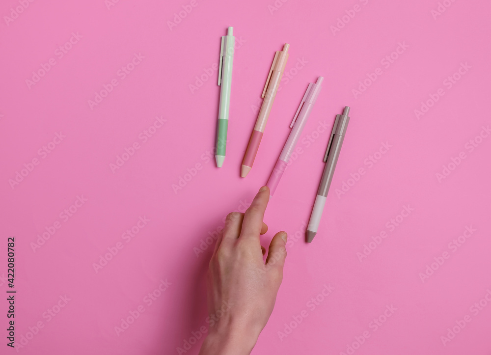 Female hand selects pen on pink background. Top view
