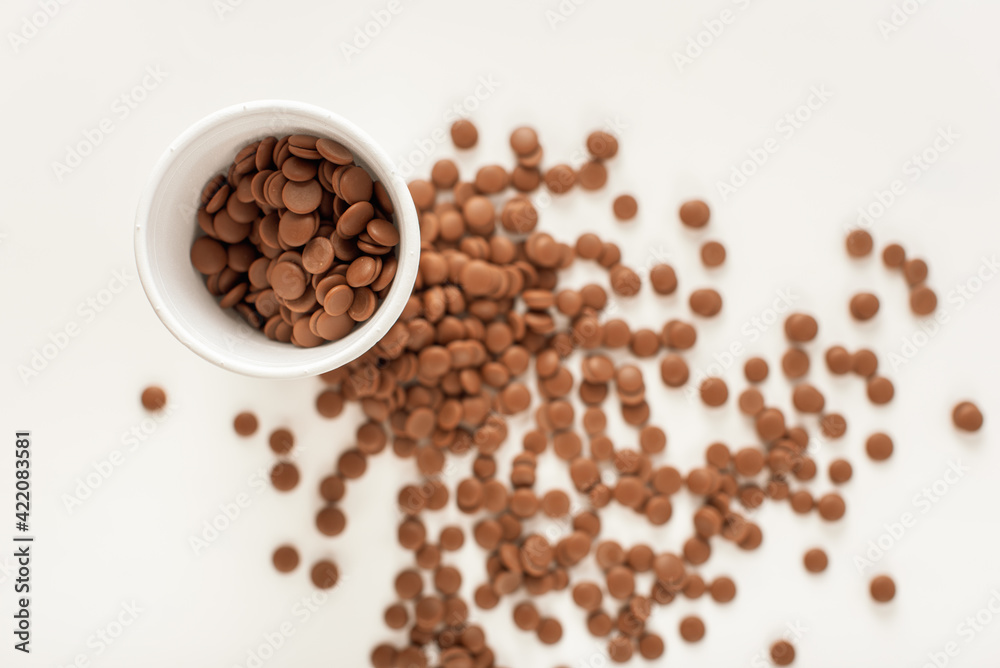 Chocolate callets in a white disposable cup on a white background. top view close-up. Flat lay. Wholesale of chocolate. Tempered chocolate confectionery concept. Chocolate callets in a white disposable cup on a white background. top view close-up. Flat lay. Wholesale of chocolate. Tempered chocolate confectionery concept.