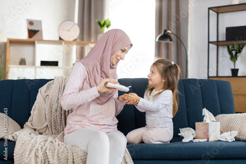 Indoor shot of lovely Muslim family doing hygiene routine at home. Young mom applying body cream or lotion on hands of little adorable daughter, looking each other