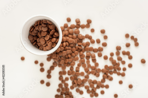 Chocolate callets in a white disposable cup on a white background. top view close-up. Flat lay. Wholesale of chocolate. Tempered chocolate confectionery concept.