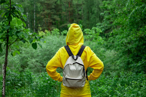 Shot back a young woman in a yellow hoodie and backpack stands in the summer forest.