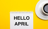 On a bright yellow background, a white cup with coffee and a white notepad with the words HELLO APRIL
