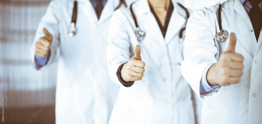 Group of unknown doctors stand as a team with thumbs up in a hospital office. Physicians ready to examine and help patients. Medical help, insurance in health care, best desease treatment and medicine Group of unknown doctors stand as a team with thumbs up in a hospital office. Physicians ready to examine and help patients. Medical help, insurance in health care, best desease treatment and medicine