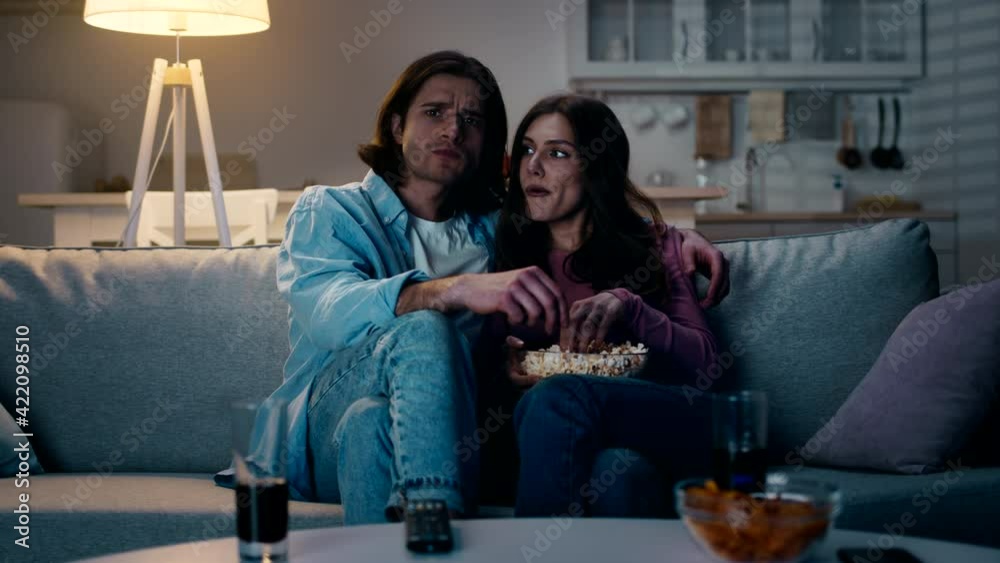 Indoors shot of young concentrated couple in love watching film with popcorn, spending evening at home, slow motion Indoors shot of young concentrated couple in love watching film with popcorn, spending evening at home, slow motion