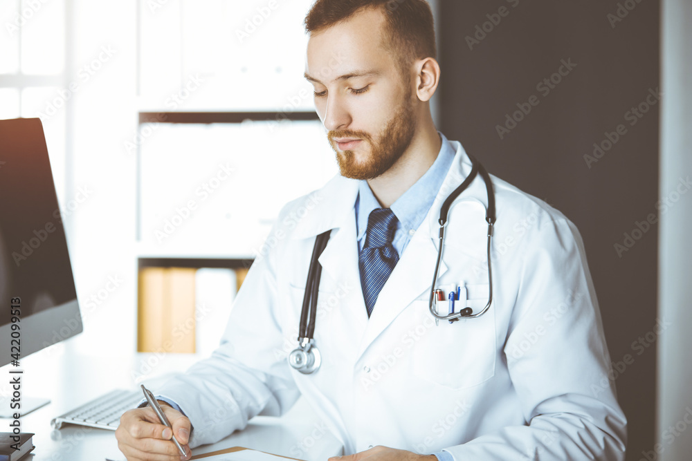 Friendly red-bearded doctor sitting and writing at clipboard in sunny clinic. Medicine concept, pandemic stop