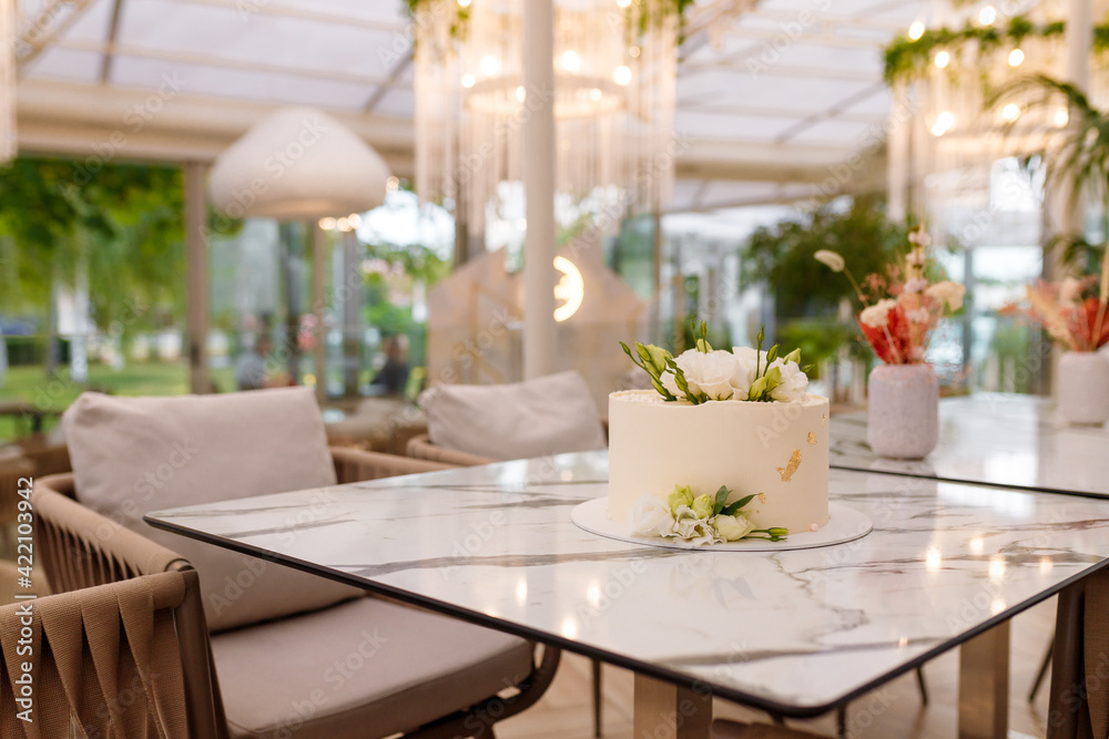 Soft focused shot of beautiful white wedding or birthday cake with roses flowers on marble table, restaurant background. Festive event sweet bakery