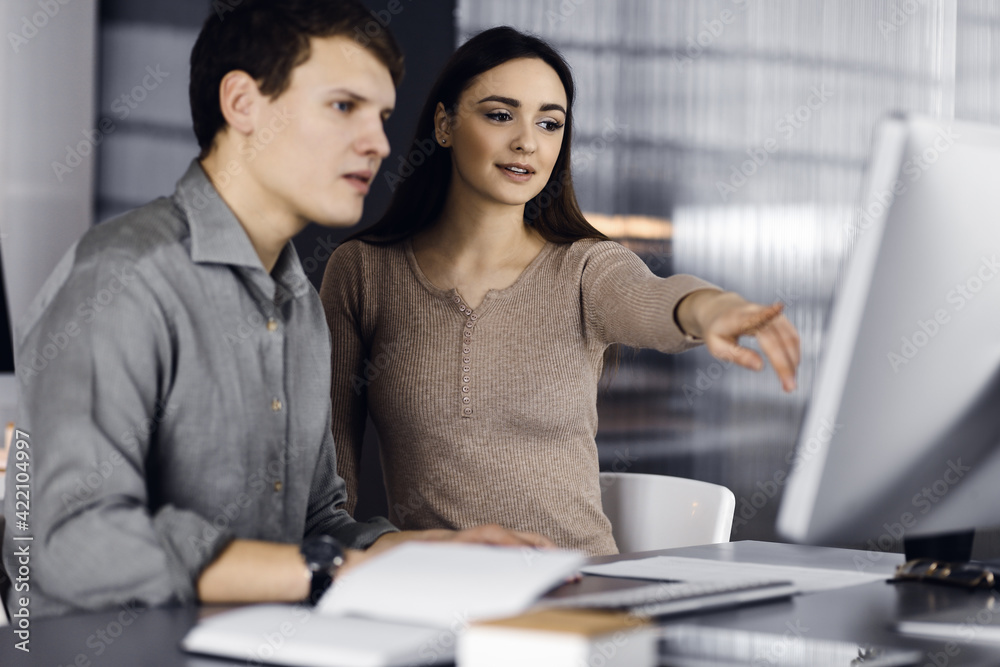 Friendly young businessman and programmer in a green shirt is working on computer, while sitting together at the desk with a female colleague in a modern office. Focus on woman. Concept of successful Friendly young businessman and programmer in a green shirt is working on computer, while sitting together at the desk with a female colleague in a modern office. Focus on woman. Concept of successful
