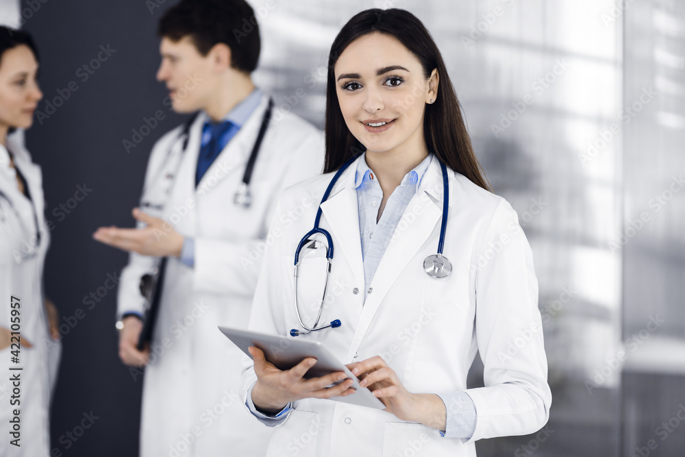 Smiling intelligent woman-doctor is holding a tablet computer in her hands, while she is standing together with her colleagues in a clinic. Physicians at work. Perfect medical service in a hospital Smiling intelligent woman-doctor is holding a tablet computer in her hands, while she is standing together with her colleagues in a clinic. Physicians at work. Perfect medical service in a hospital