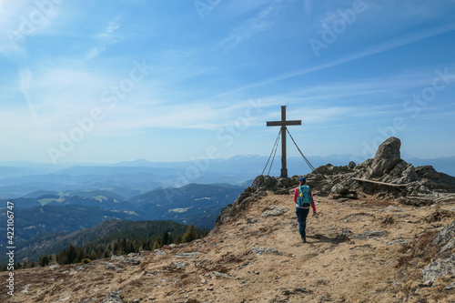 A woman with hiking backpack reaching the top of Sauofen in Austrian Alps. A wooden cross on top. Lots of stones around. Fall vibes. High mountain chains in the back. Serenity and achievement photo