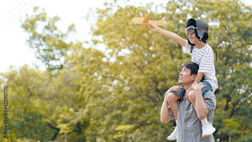 Lifestyle Asian family Father and son sat on his shoulders and run in park. Paper plane as a toy in hand of kid And there are many big trees in garden Is natural in the morning of summer