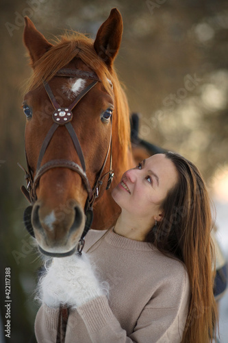 Close portrait of young smiling woman and brown horse at winter sunset. Woman with long hear in sweater near big horse