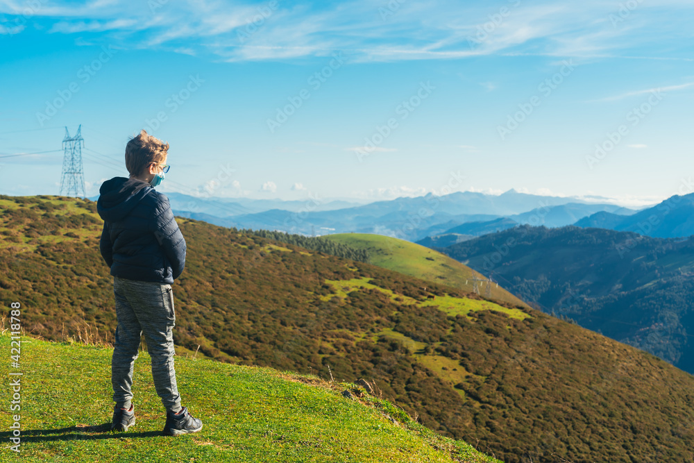 blond boy wearing glasses and protected by a covid mask, looks at the horizon on the green mountain 