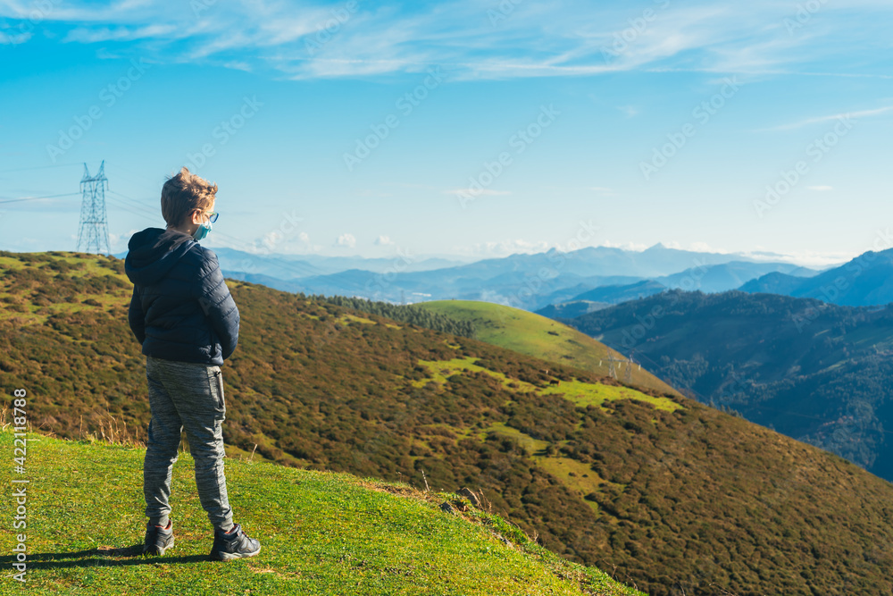 blond boy wearing glasses and protected by a covid mask, looks at the horizon on the green mountain blond boy wearing glasses and protected by a covid mask, looks at the horizon on the green mountain