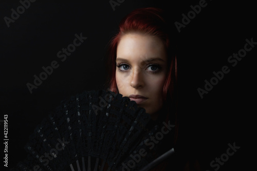 Low key portrait of beautiful young woman hiding her face behind the fan. Horizontally. 