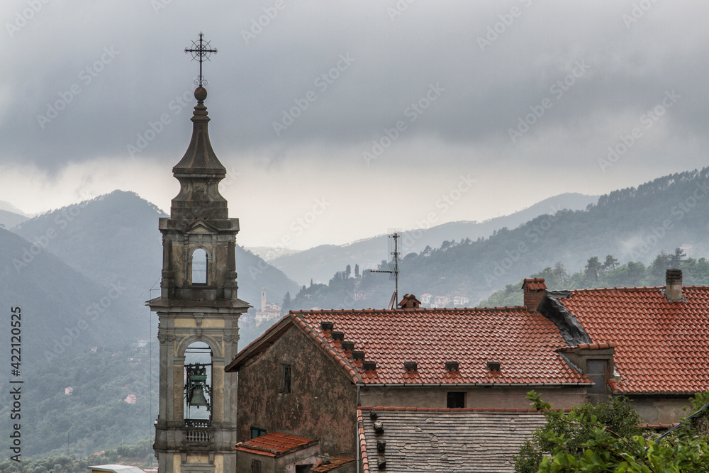 Obraz premium The bell tower of the temple against the background of mountains, surrounded by houses with tiled roofs. Low clouds after rain.
