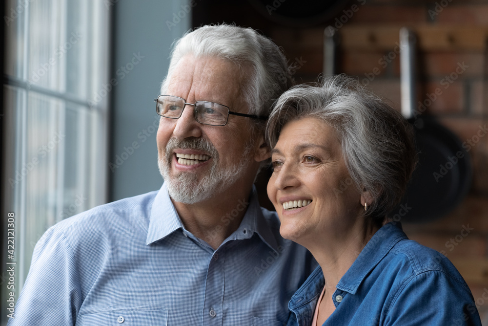 Lost in dreams. Headshot of affectionate elderly married couple pensioners cuddle look away with happy white smiles enjoy love care create plans. Happy healthy aged spouses stand by window think dream Lost in dreams. Headshot of affectionate elderly married couple pensioners cuddle look away with happy white smiles enjoy love care create plans. Happy healthy aged spouses stand by window think dream