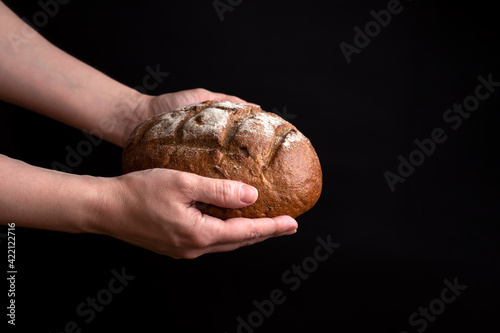 Large round loaf of rye bread in female hands on a black background.