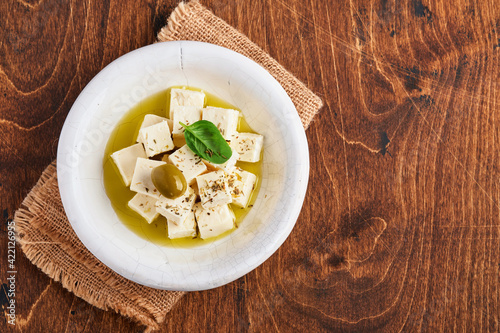 Feta cheese cubes with rosemary, olives and olive oil sauce in white bowl on old brown wooden background. Traditional Greek homemade cheese. Selective focus.