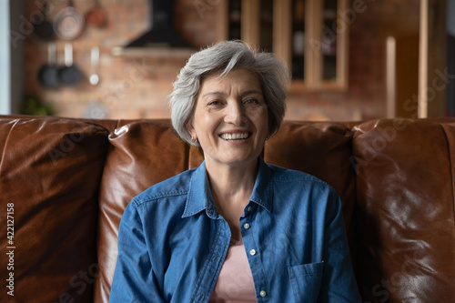 Active old age. Portrait of optimistic retired hispanic female clothed in stylish casual demonstrating perfect white teeth in beautiful smile. Energetic senior latin woman sit on couch look at camera