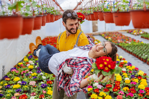 Cheerful couple in love having fun at greenhouse. Man carrying woman while woman holding pot with flowers. © dusanpetkovic1