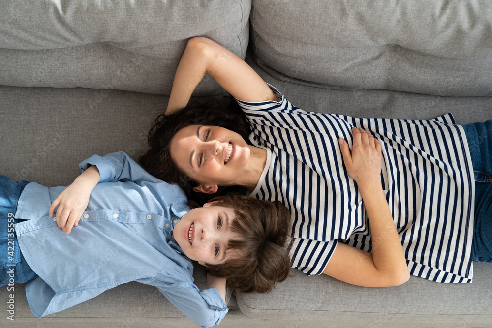 Top above view of happy young mom and little kid son relaxing on sofa at home. Couple of carefree mother and cute child lying, resting on couch, having fun, looking together at camera. Family, love. Top above view of happy young mom and little kid son relaxing on sofa at home. Couple of carefree mother and cute child lying, resting on couch, having fun, looking together at camera. Family, love.