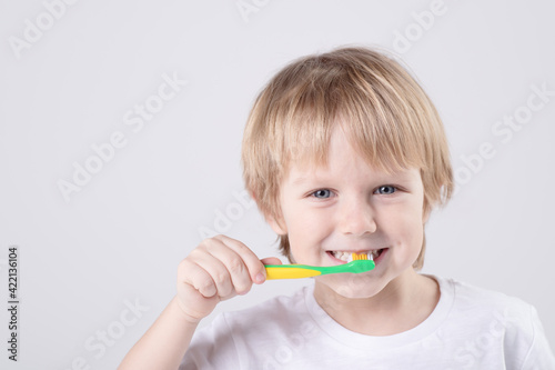 Funny Caucasian child brushes teeth. Eye contact. Studio light. White T-shirt. Concept of baby teeth, healthy lifestyle, routine, development of a child's skill to brush his own teeth. Copy space
