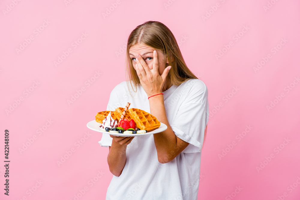 Young russian woman eating a waffle isolated blink through fingers frightened and nervous.