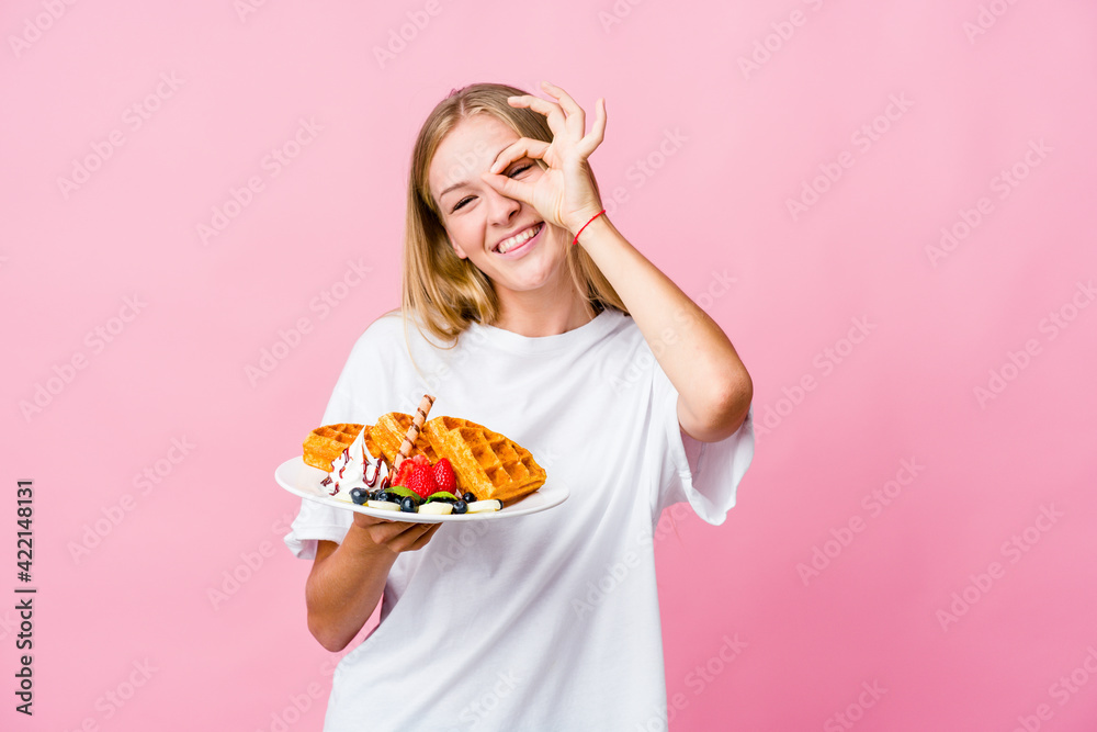 Young russian woman eating a waffle isolated excited keeping ok gesture on eye.