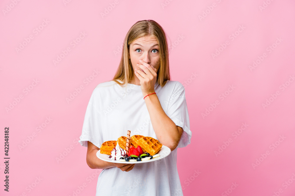 Young russian woman eating a waffle isolated shocked covering mouth with hands.