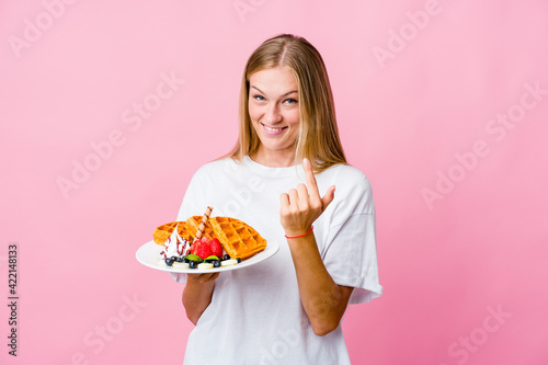 Young russian woman eating a waffle isolated pointing with finger at you as if inviting come closer.