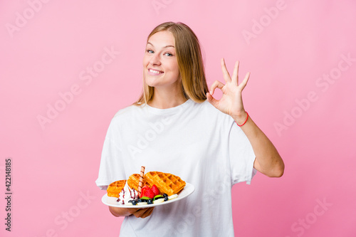 Young russian woman eating a waffle isolated cheerful and confident showing ok gesture.