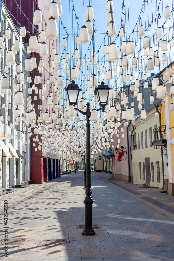 Obraz premium Decorations of Stoleshnikov lane in Moscow. Large garland from electric bulbs hangs above pedestrian street. Clear blue sky in the background. Travel in Russia theme.